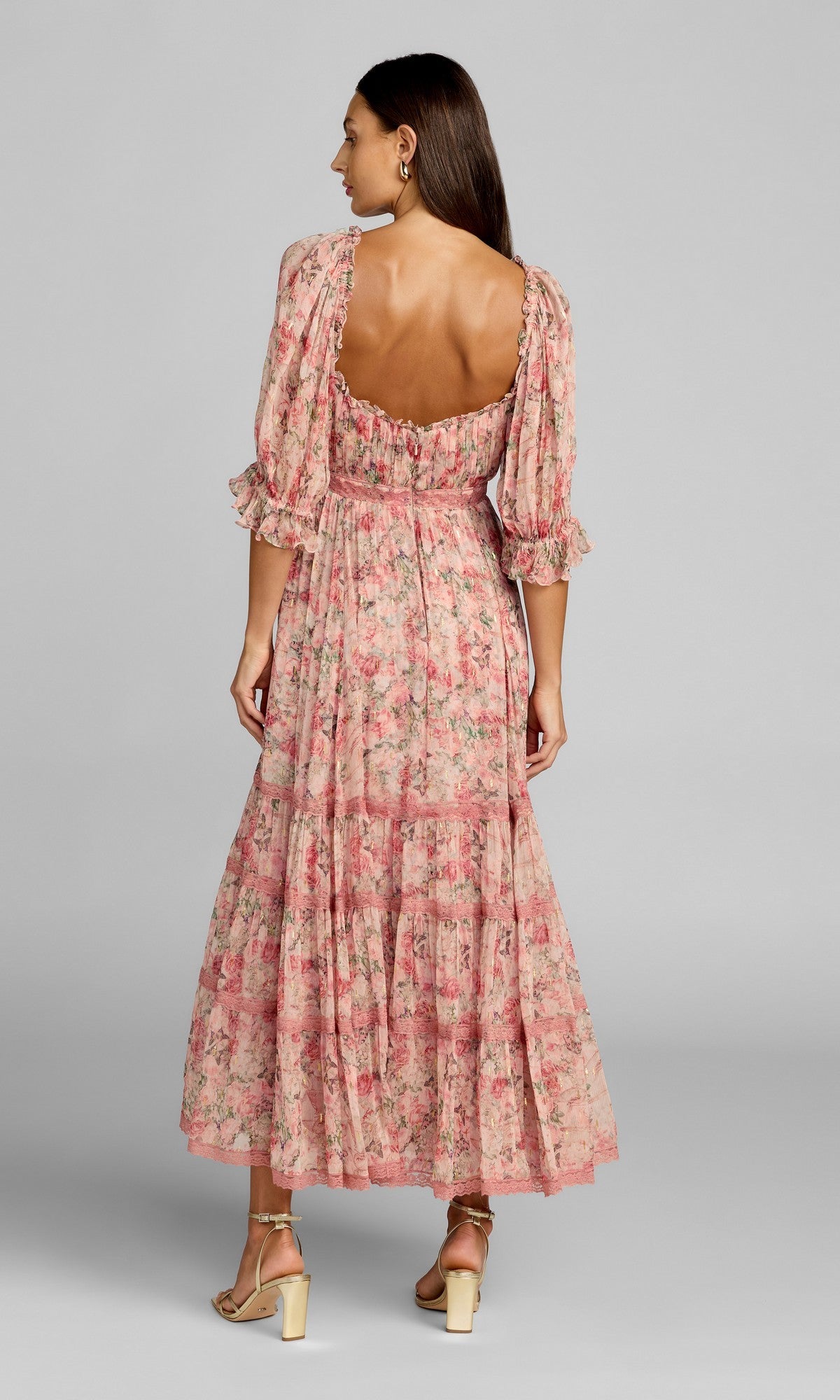 A woman stands facing away, wearing the Mac Duggal 8330 tea-length party dress—a pink floral design with puff sleeves and ruffled trim—paired with light-colored heeled sandals against a plain background.